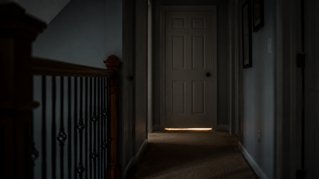 A dark, generic upstairs hallway with a closed bedroom door, soft light under the door, no faces, no text.